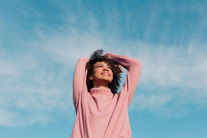 Jeune femme heureuse portantun col roulé rose, souriante au soleil, contre un ciel bleu lumineux enarrière-plan