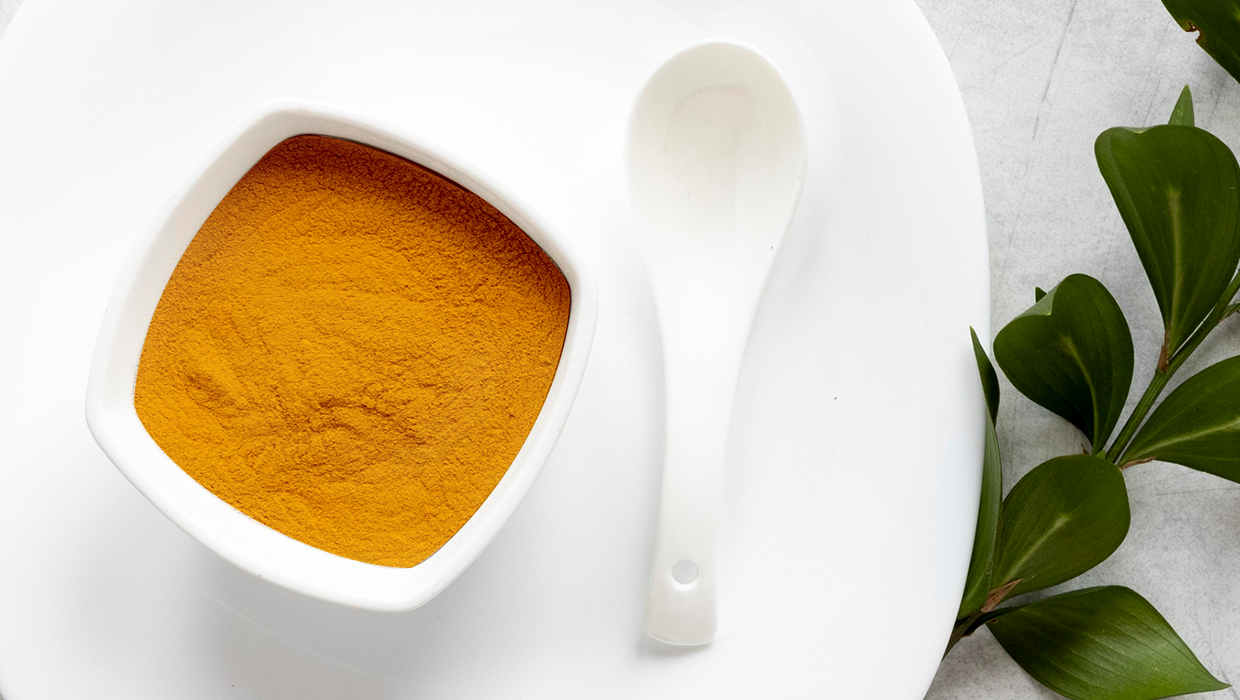 A white bowl filled with bright yellow turmeric powder, accompanied by a white spoon, on a light surface with green leaves nearby.