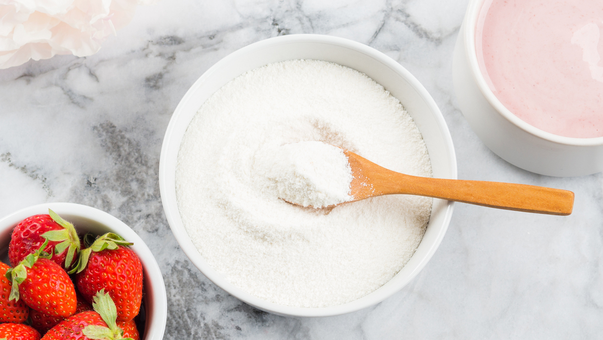 A bowl of sugar with a wooden spoon, fresh strawberries in a nearby bowl, and a small dish of pink yogurt on a marble surface.
