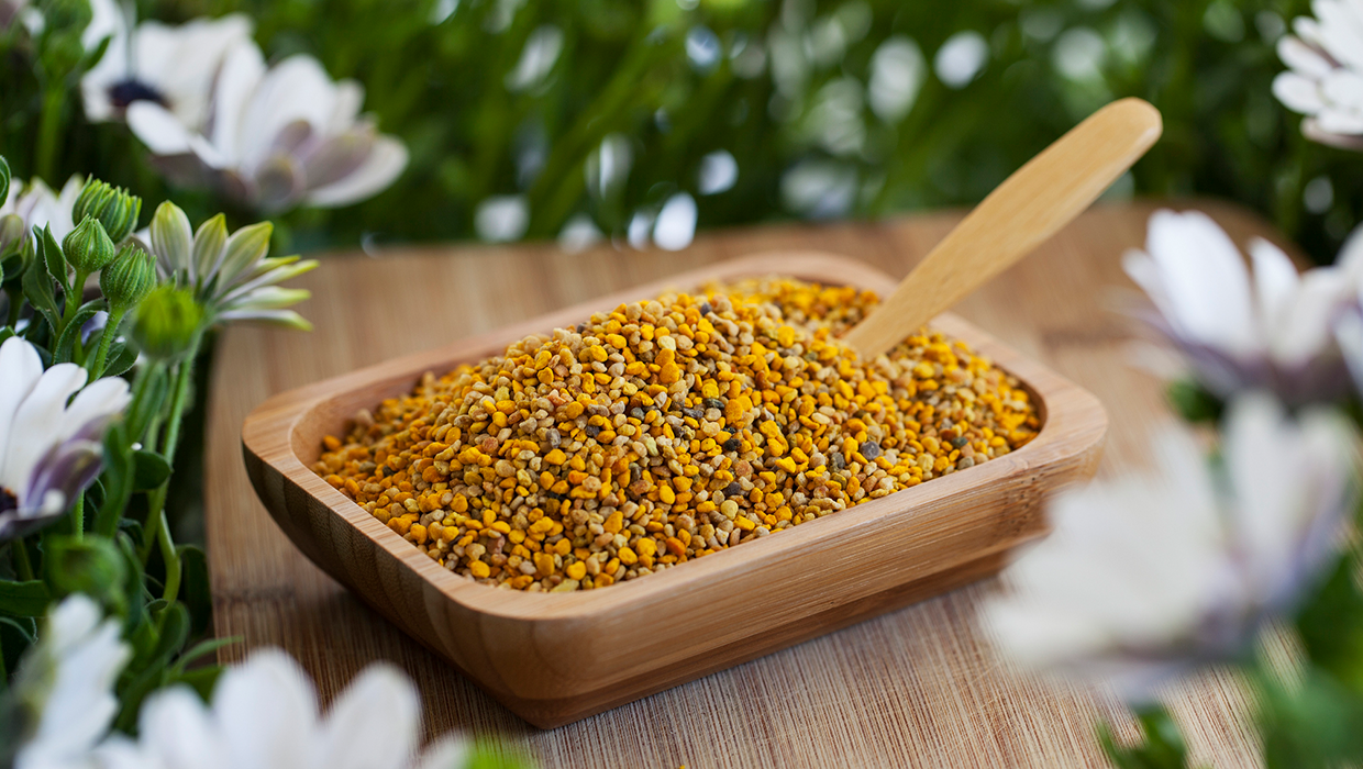 A wooden bowl filled with colorful pollen grains, surrounded by green foliage and white flowers.