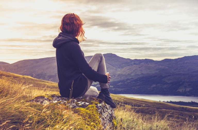 Red haired woman sits serenely on a hill overlooking a mountaintop lake