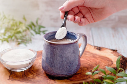 A hand holds a spoonful of powder above a mug of coffee, with a small bowl of powder nearby, set on a wooden surface.