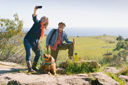 A woman and an older man take a selfie outdoors with a golden retriever, surrounded by greenery and a scenic view.