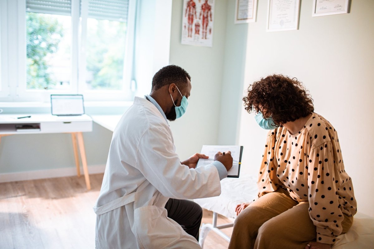 A doctor in a white coat is consulting with a patient wearing a polka dot top in a medical office. Both are wearing masks.