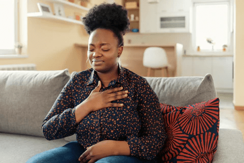 A woman sits on a couch, holding her chest with a thoughtful expression, in a cozy living room setting.