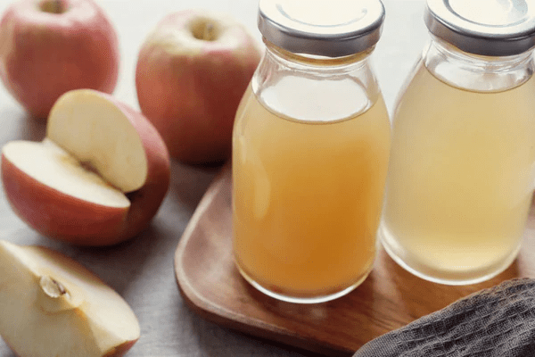 Two glass bottles of apple juice on a wooden tray, surrounded by whole and sliced apples.