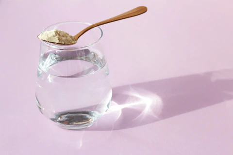 A glass of water with a spoonful of powder resting on the rim, set against a soft pink background.