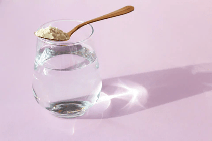 A glass of water with a spoonful of powder resting on the rim, set against a soft pink background.