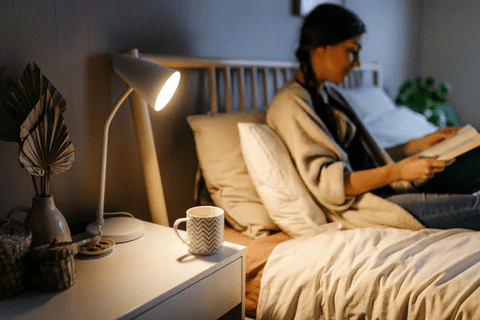 A cozy bedroom scene with a woman reading on a bed, a lamp illuminating the space, and a mug on a nightstand.