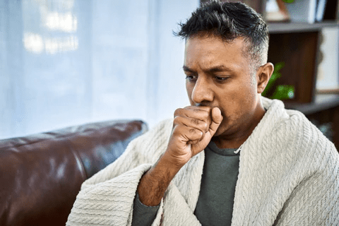 A man wrapped in a blanket sits on a couch, looking concerned while coughing. Natural light filters through a window.