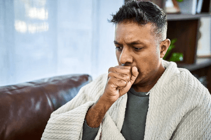A man wrapped in a blanket sits on a couch, looking concerned while coughing. Natural light filters through a window.