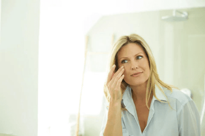 A woman with long blonde hair is applying skincare in a bright bathroom mirror. She looks thoughtful and relaxed.